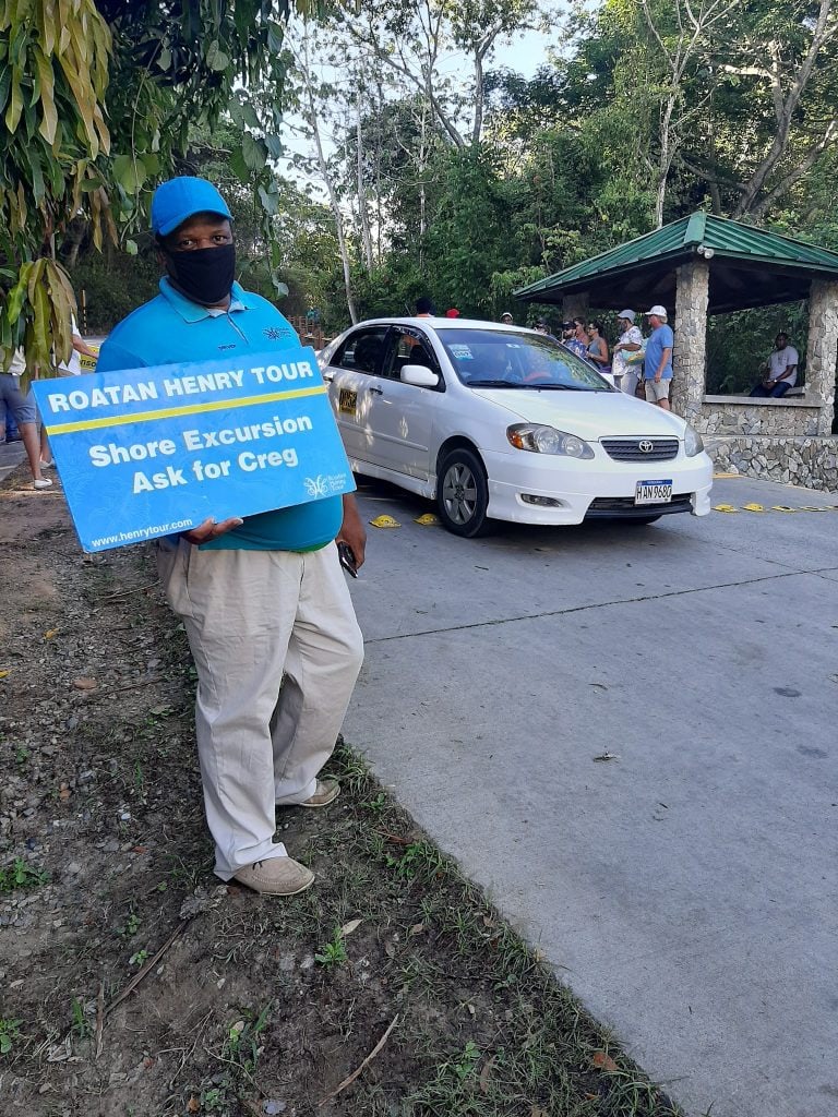 A man wearing a blue cap, black face mask, and a blue shirt holding a sign that reads "Roatan Henry Tour Shore Excursion Ask for Creg" stands beside a white taxi on a paved road surrounded by greenery and trees in Roatan, Honduras.