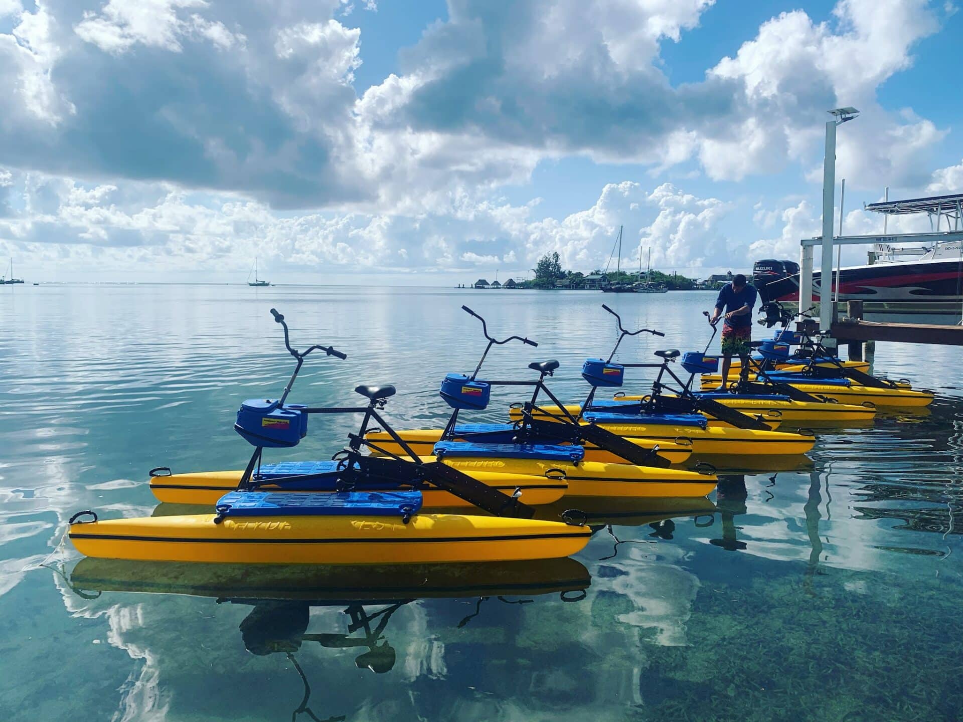 Yellow paddle boats are docked along the calm waterfront in Roatan, Honduras, with sailboats and a small island with overwater bavilions visible in the background under a partly cloudy sky.