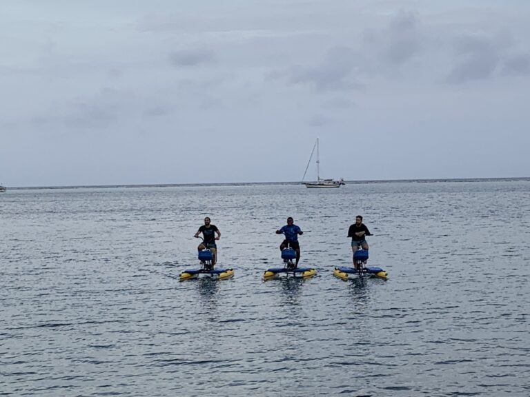 Three people are paddleboarding on the ocean near Roatan, Honduras, with a sailboat in the distance under a cloudy sky.