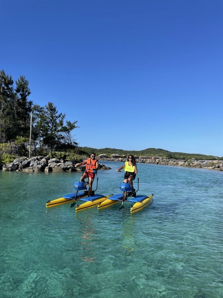 Two people are riding yellow paddleboards with electric motors in clear turquoise waters near Roatan, Honduras, with a rocky shoreline, lush green trees, and a bright blue sky in the background.