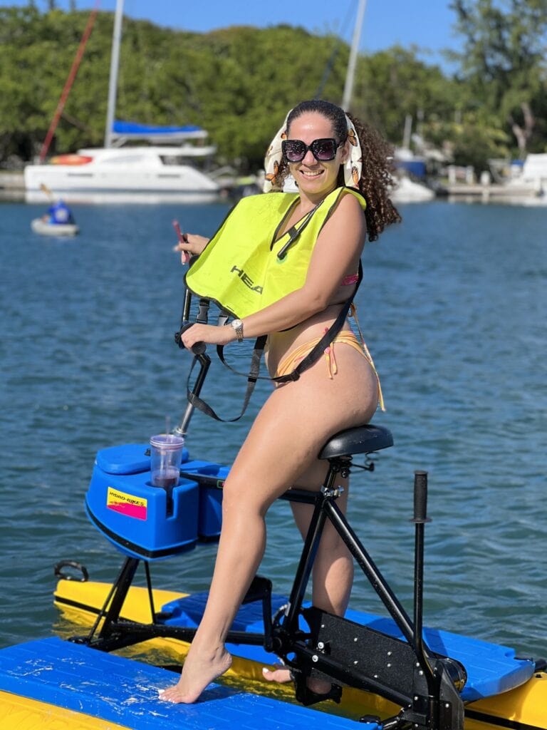 A woman in a bikini and sunglasses pedals a yellow and blue paddleboard with a mounted electric motor on Roatan, Honduras, with sailboats and lush green trees in the background.