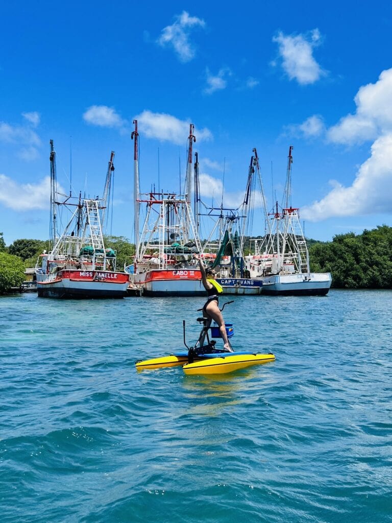A person riding a yellow stand-up paddleboard on the water near a marina with several sailboats docked under a bright blue sky in Roatan, Honduras.