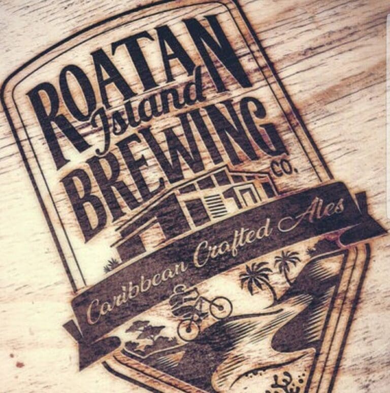A close-up of a wooden sign engraved with the logo of Roatan Brewing Co., showcasing a mountain and palm tree design, on a light wooden surface, representing a brewery located in Roatan, Bay Islands, Honduras.