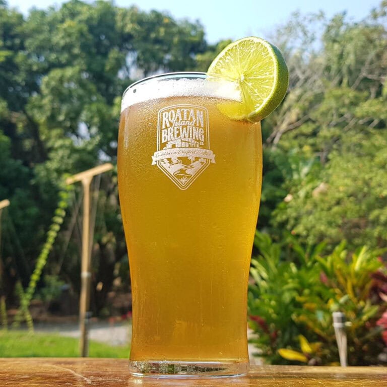 A tall glass of cold beer with a lime wedge garnished on the rim, featuring the Roatan Brewing logo, is set outdoors against a lush green tropical background in Roatan, Honduras.