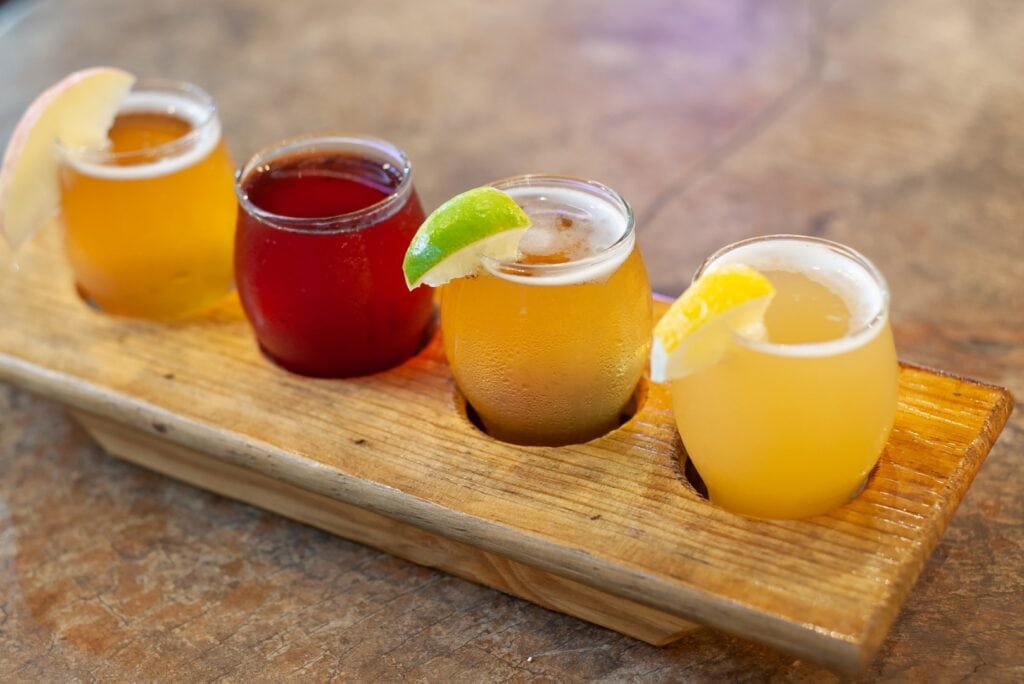 A wooden serving board holds four colorful cocktails with lemon and lime garnishes, set on a rustic table in Roatan, Honduras.
