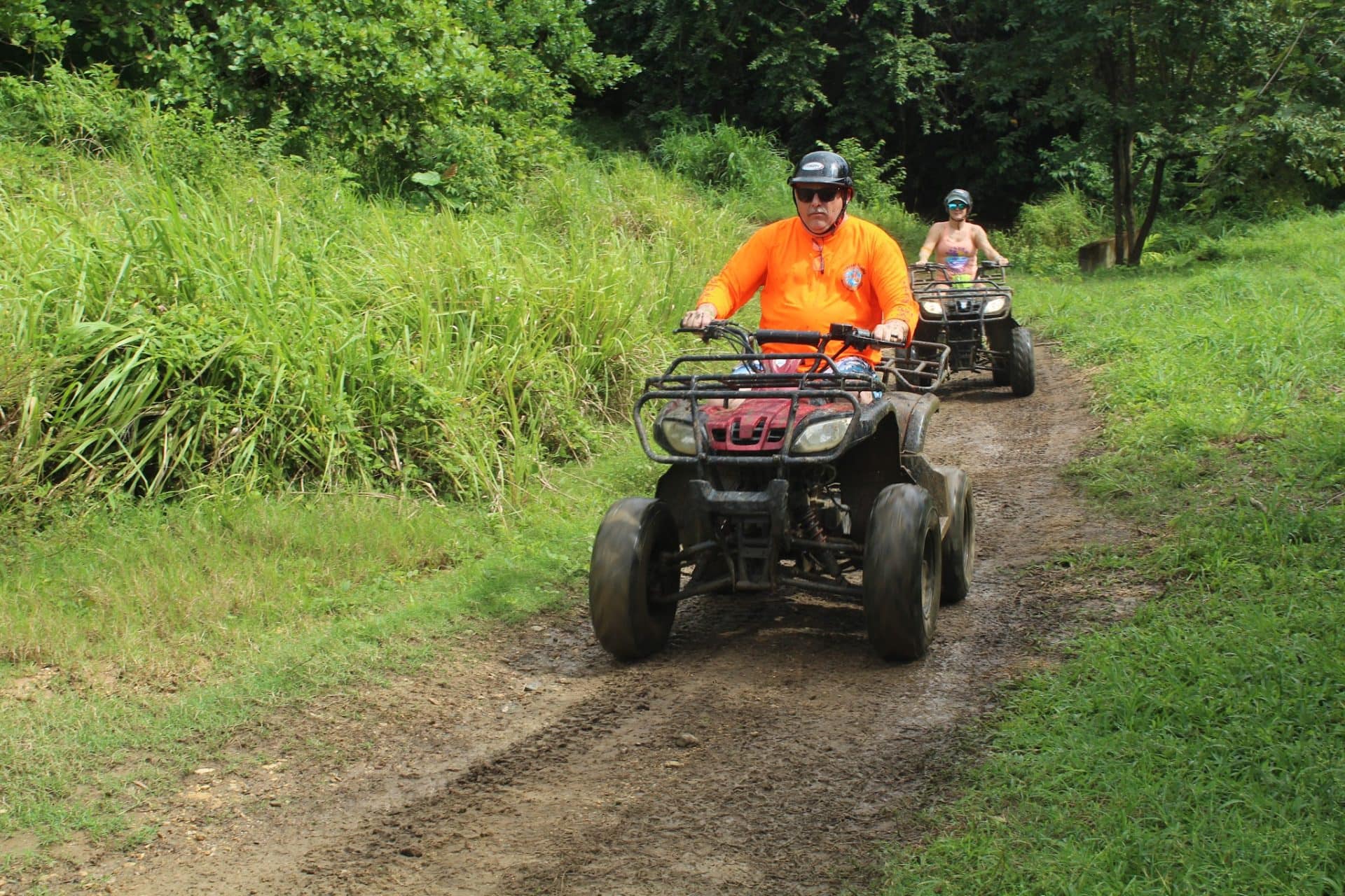 Two people riding all-terrain vehicles along a narrow dirt trail surrounded by lush green vegetation in Roatan, Honduras.