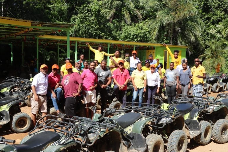 A group of people in colorful clothing, including some wearing orange caps, pose with all-terrain vehicles in front of a green and yellow structure surrounded by lush tropical trees on Roatan, Honduras.