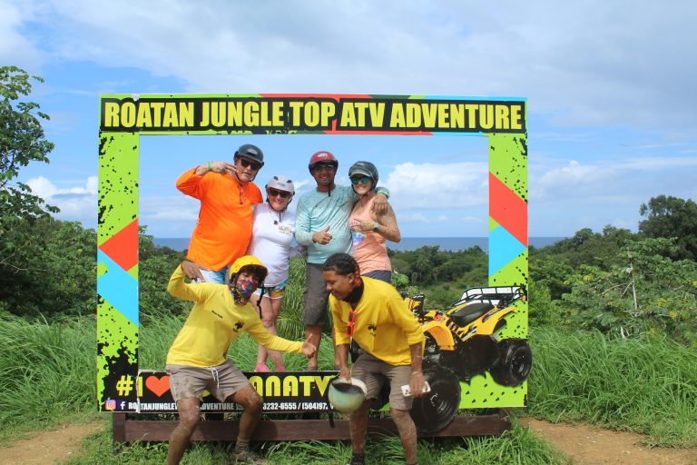 A group of people in colorful outdoor gear pose under a sign for Roatan Jungle Top ATV Adventure, with lush green vegetation and a clear blue sky in the background on Roatan, Honduras.