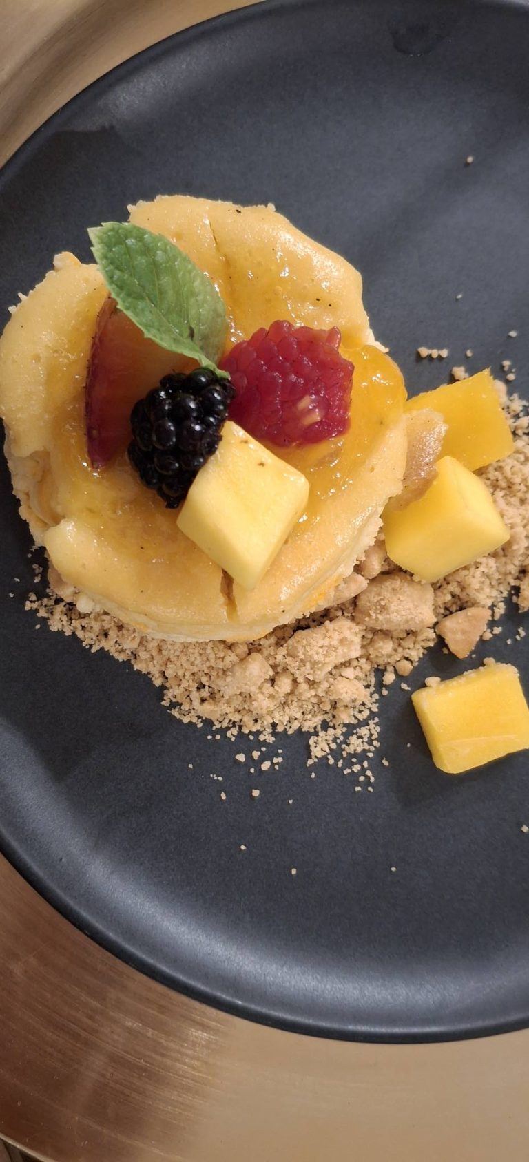 A plated dessert featuring mango chunks, fresh blackberries, raspberries, and a sprig of mint on a bed of crushed cookie crumbs, served on a dark plate in Roatan, Honduras.
