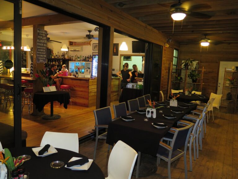 The image shows the interior of a restaurant or bar in Roatan, Honduras, featuring wooden walls and ceiling, with tables set for dining, some decorated with flowers, and a bar area with bottles and a bartender preparing drinks.