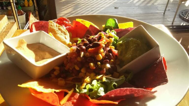 A colorful plate of fresh vegetables, black bean salad, and crispy plantain chips is displayed on a white dish at a restaurant in Roatan, Honduras, with small bowls of dip or sauce and vibrant side garnishes.