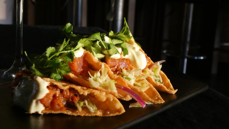A plate of crispy tacos topped with shredded lettuce, cilantro, sour cream, and sauce is displayed on a black tray in a dimly lit setting on Roatan, Honduras.
