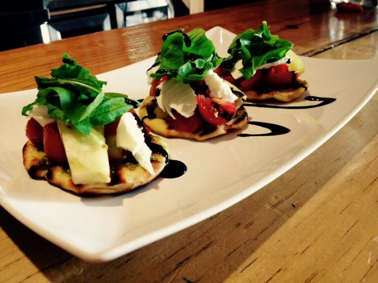 Wood-fired mini pizzas topped with fresh greens, cherry tomatoes, cheese, and balsamic glaze are displayed on a white rectangular plate in a restaurant setting in Roatan, Bay Islands, Honduras.
