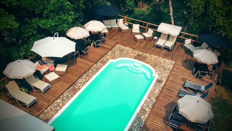 A wooden deck surrounds a rectangular swimming pool with lounge chairs, umbrellas, and seating areas, set amidst lush greenery in Roatan, Honduras.
