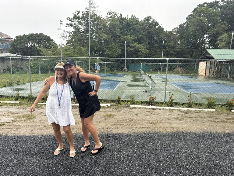 Two women stand smiling together on an overcast day near a fenced tennis court in Roatan, Honduras, with lush green trees and a building with a green roof visible in the background.