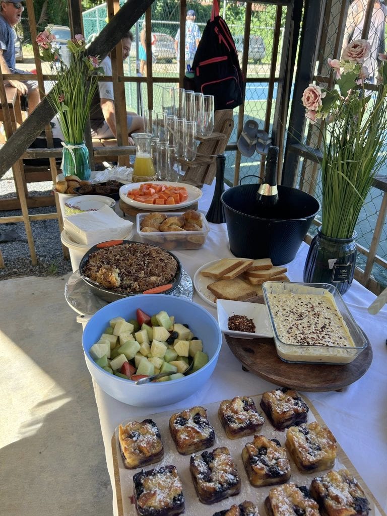 A spread of breakfast foods including fruit, baked goods, and desserts is displayed on a table outdoors at a business in Roatan, Honduras, with people sitting nearby and a tennis court visible in the background.