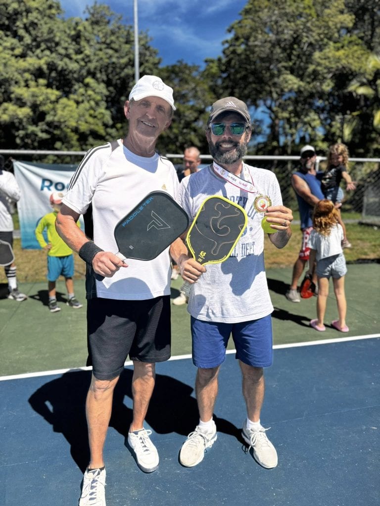 Two men stand together on a tennis court in Roatan, Honduras, smiling and holding pickleball paddles, with other people and trees visible in the background.