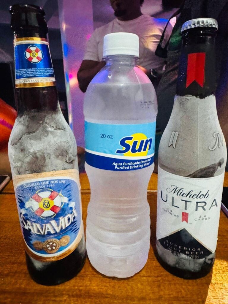 Three bottles of alcoholic and non-alcoholic beverages, including Salva Vida beer, a 20 oz bottle of Sun purified drinking water, and Michelob Ultra beer, are displayed on a wooden table with a blurred background featuring people and colorful lighting, in Roatan, Honduras.
