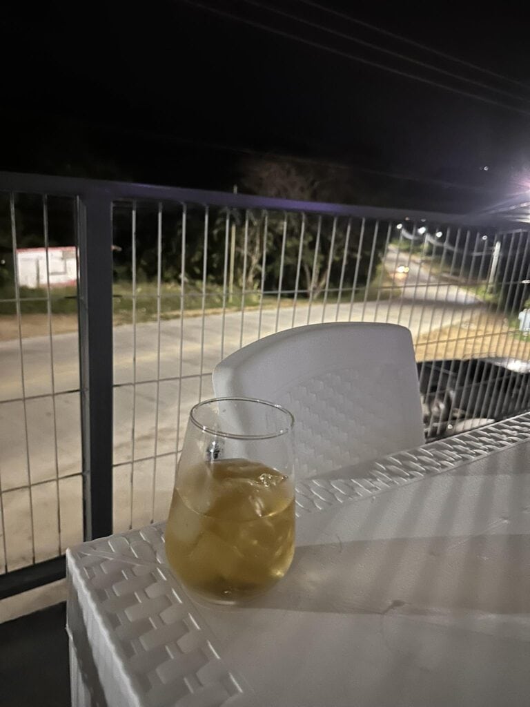 A glass of iced tea sits on a white plastic table outside an establishment in Roatan, Honduras, with a black metal fence and a street with trees and streetlights visible in the background at night.