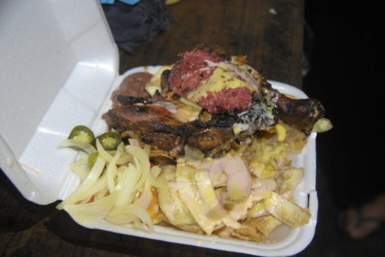 A takeout container in Roatan, Honduras, holds a serving of loaded fries topped with grilled chicken, melted cheese, and a generous portion of flaky, breaded fish fillet with a dollop of sauce and a side of pickled jalapeños and sliced onions.