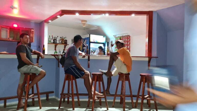 Three men are sitting on wooden stools at a bar in a casual interior within Roatan, Honduras, with a blue wall, bottles of liquor, and a refrigerator visible behind the bar, as part of a lively social scene.