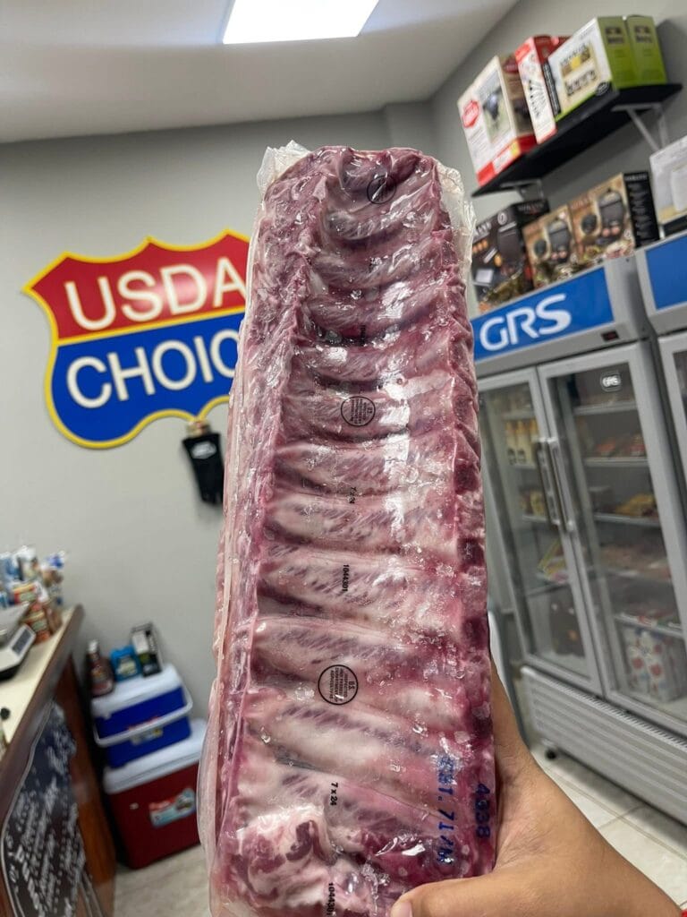 A hand holding a large sealed package of raw beef in a grocery store aisle with a "USDA Choice" sign, refrigeration units, and store shelves visible in the background in Roatan, Honduras.