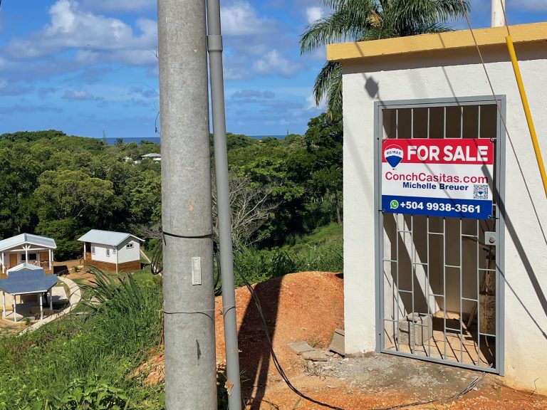 A white building with a "For Sale" sign from ConchCasitas.com and part of a yellow roof is situated in a lush, green, tropical area on Roatan in Honduras, with palm trees and a view of other small houses and the ocean in the background.