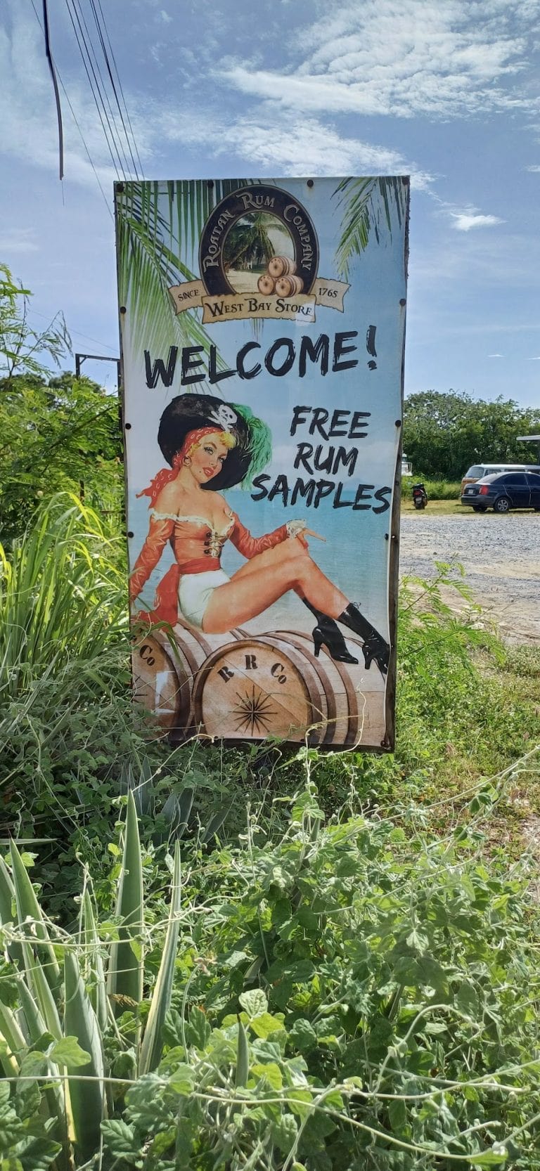 A colorful outdoor sign in Roatan, Honduras, advertises free rum samples with an illustration of a woman dressed in vintage-inspired clothing sitting on a wooden barrel, surrounded by lush green foliage, under a partly cloudy sky.