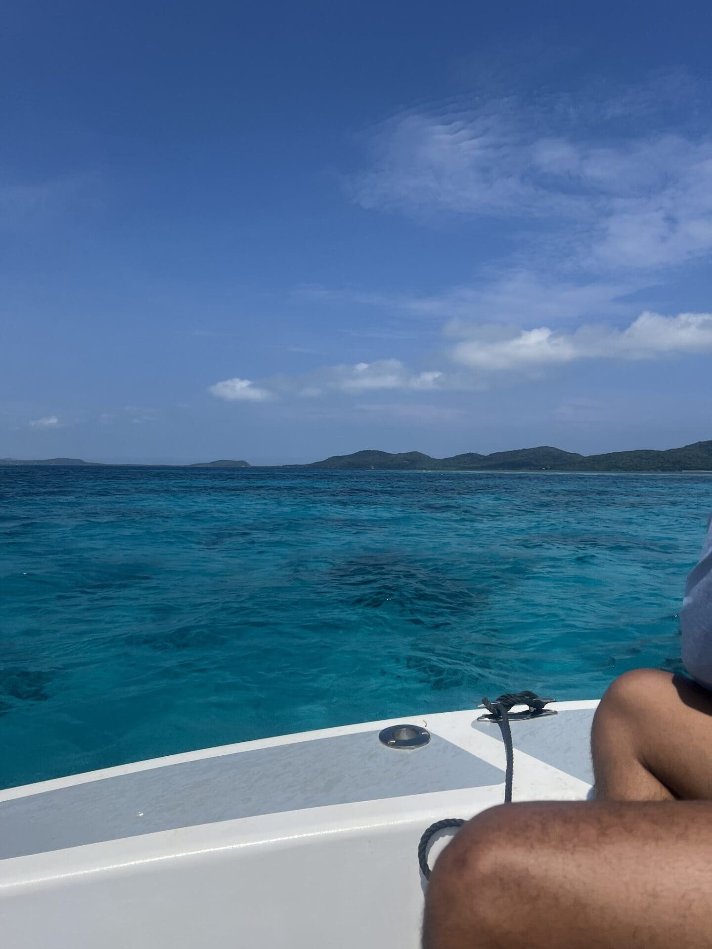 A person’s tanned arm is seen on a boat deck with turquoise waters and distant green islands under a partly cloudy sky in Roatan, Honduras.