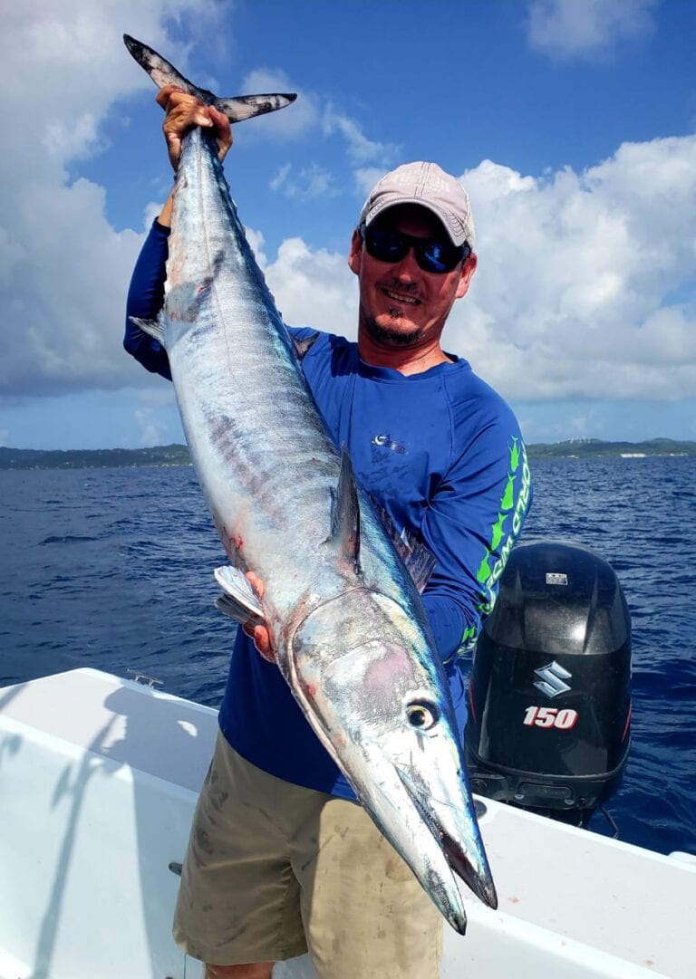 A man on a boat in Roatan, Honduras, holds up a large fish he caught while fishing offshore, with the ocean and partly cloudy sky in the background.