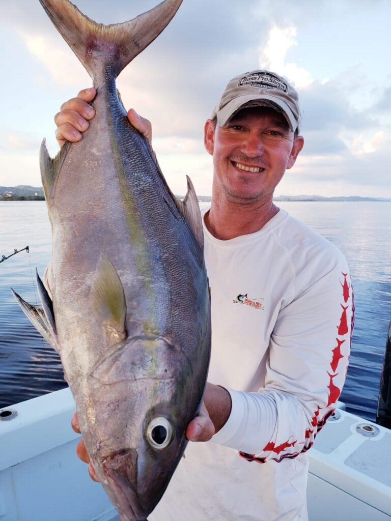 A man smiling on a boat in Roatan, Honduras, holding a large fish he caught with a scenic ocean and cloudy sky in the background.