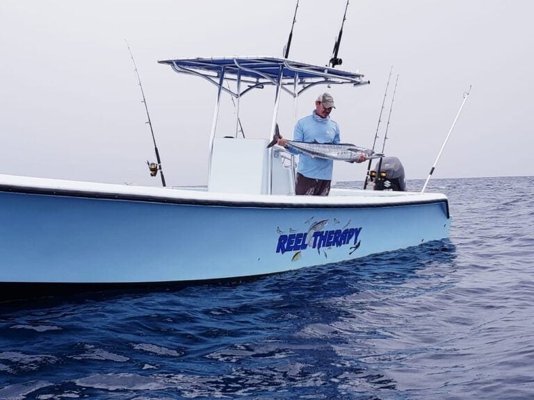 A man on a blue fishing boat named "Reel Therapy" holding a large fish, with multiple fishing rods mounted on the boat, on the open waters near Roatan, Honduras.
