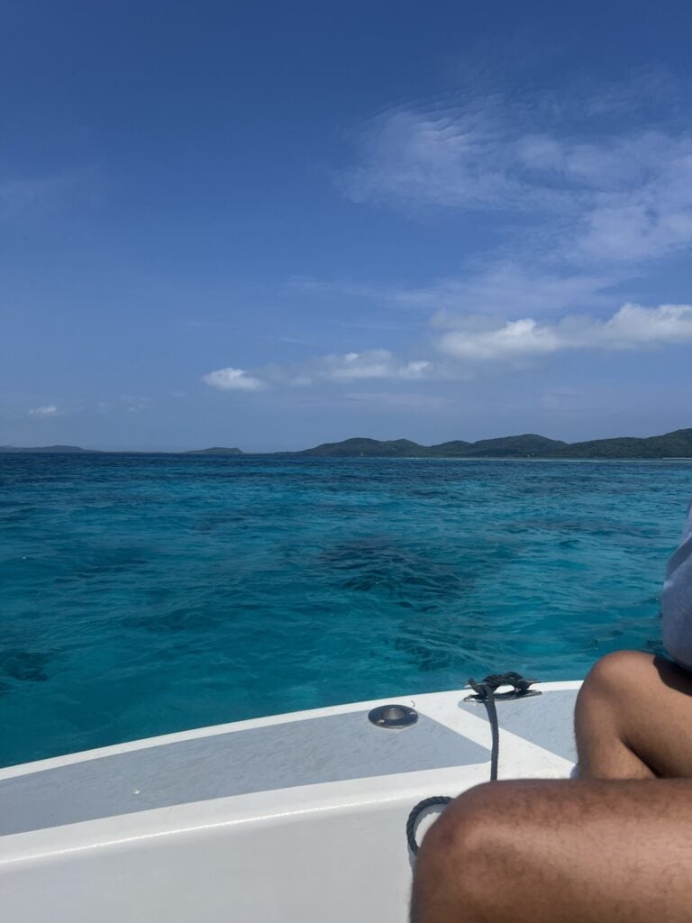A person’s tanned arm is seen on a boat deck with turquoise waters and distant green islands under a partly cloudy sky in Roatan, Honduras.