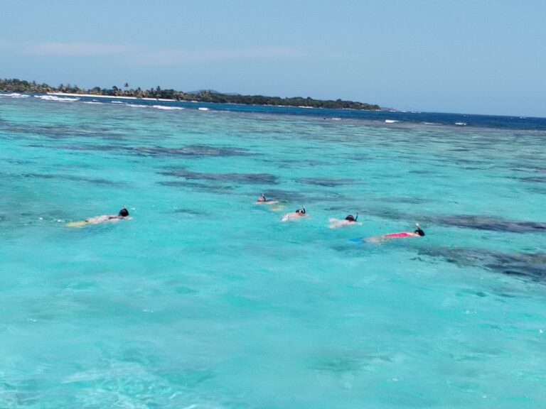 People snorkeling in clear turquoise waters near Roatan, Honduras, with a lush, green island and blue sky in the background.