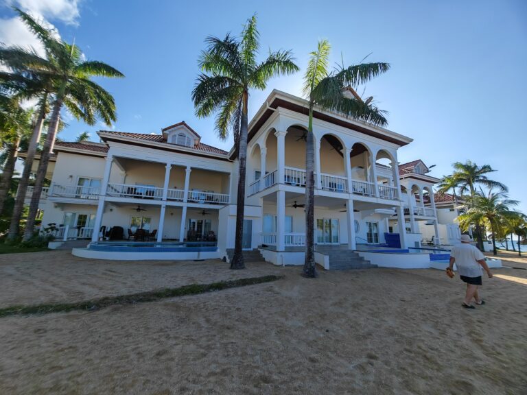 A beige multi-story building with arched balconies, surrounded by tall palm trees under a clear blue sky in Roatan, Honduras.
