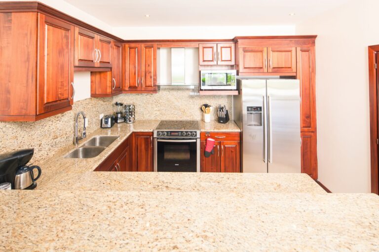 The image shows a modern kitchen with wooden cabinets, a granite countertop, and stainless steel appliances, located in Roatan, Honduras.