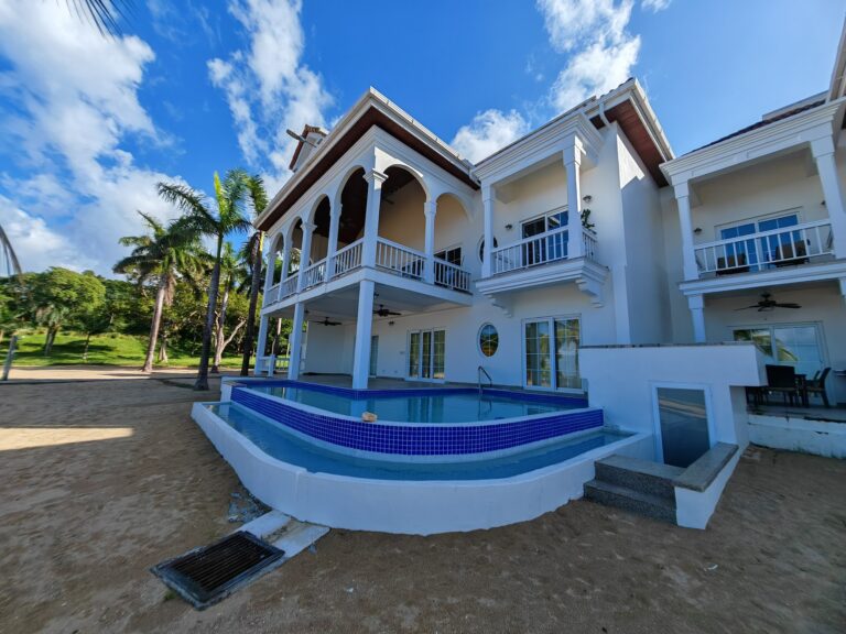 A white multi-story building with arched balconies and a swimming pool in the foreground is set against a bright blue sky with scattered clouds in Roatan, Honduras.