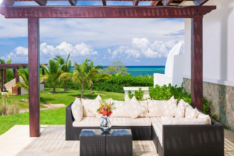 A outdoor seating area with a black wicker sectional sofa decorated with white cushions and a flower arrangement on a glass-topped coffee table, overlooking a lush green garden, palm trees, and the ocean in Roatan, Honduras.
