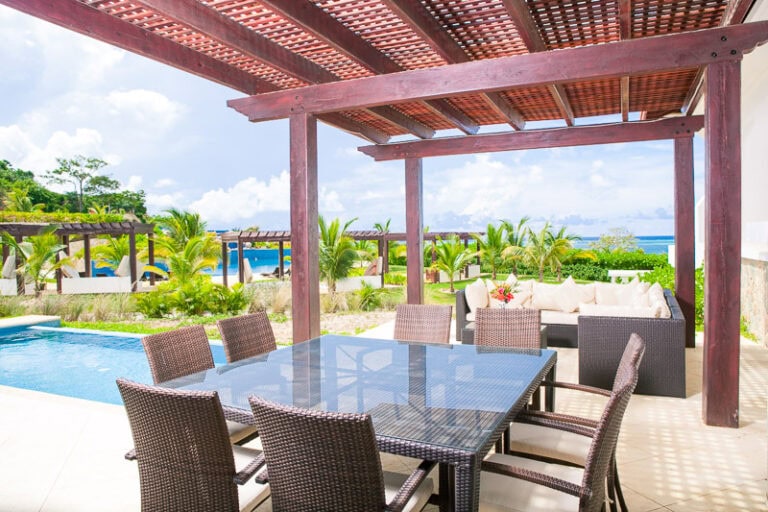 A shaded outdoor dining area on a tropical property in Roatan, Honduras, featuring a glass-topped table with six rattan chairs, a nearby swimming pool, lush palm trees, and a view of the ocean in the background.