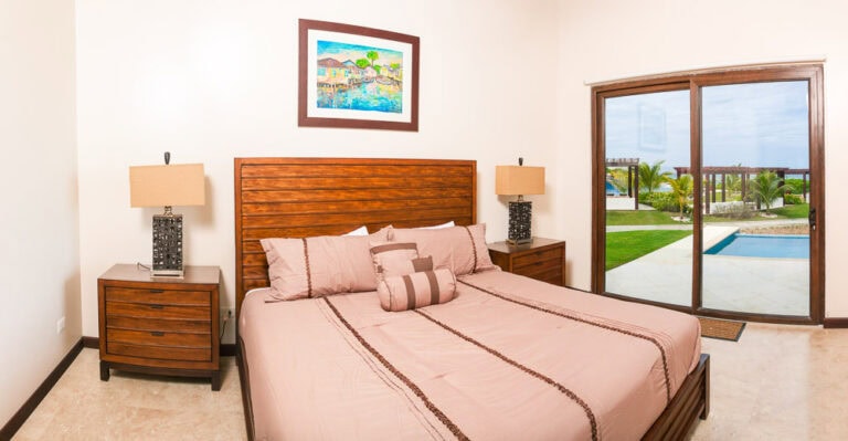 A bedroom in Roatan, Honduras, features a wooden bed with pink bedding and pillows, matching wooden nightstands with lamps, and a large sliding glass door opening to a patio with a view of a swimming pool, tropical palm trees, and the ocean.