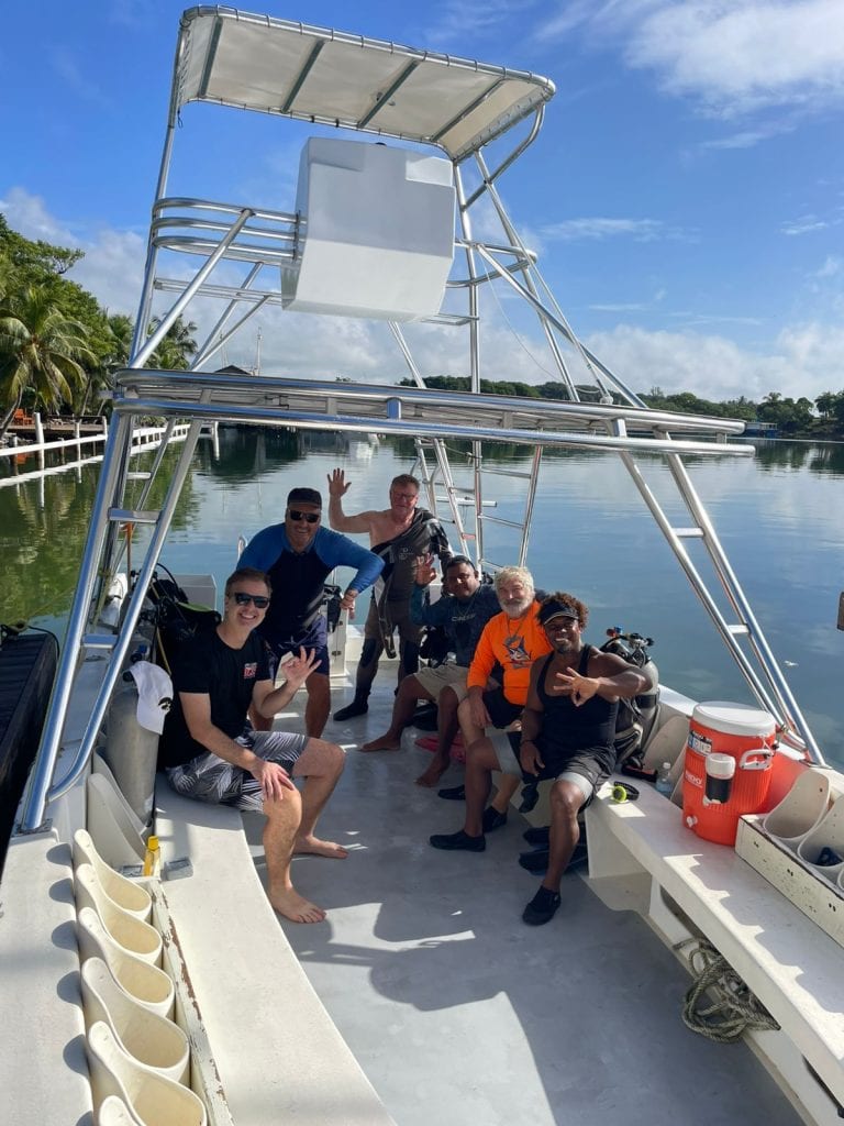 A group of six men enjoying a boat ride on calm waters in Roatan, Honduras, with lush green trees and a clear blue sky in the background.