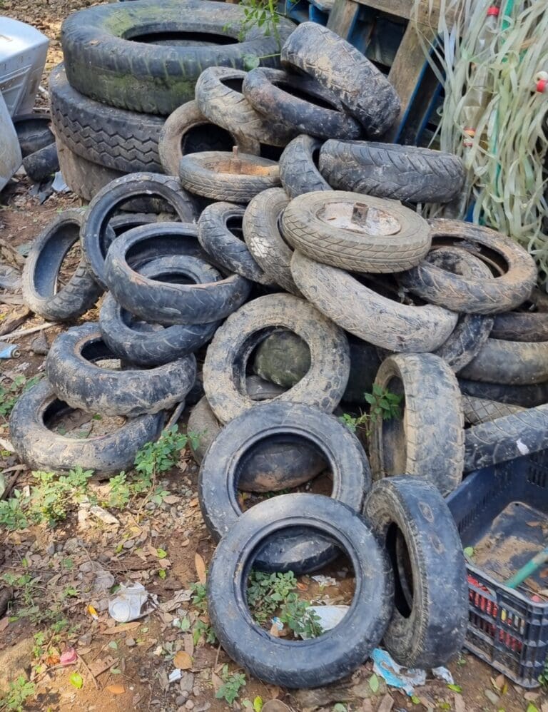 A large pile of used, weathered tires is stored outdoors on Roatan, Honduras, with some stacked on top of each other and others scattered on the ground surrounded by dirt and small plants.