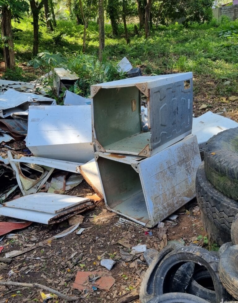 Discarded metal washing machine parts lying among tires and debris in a green, wooded area on Roatan in Honduras.