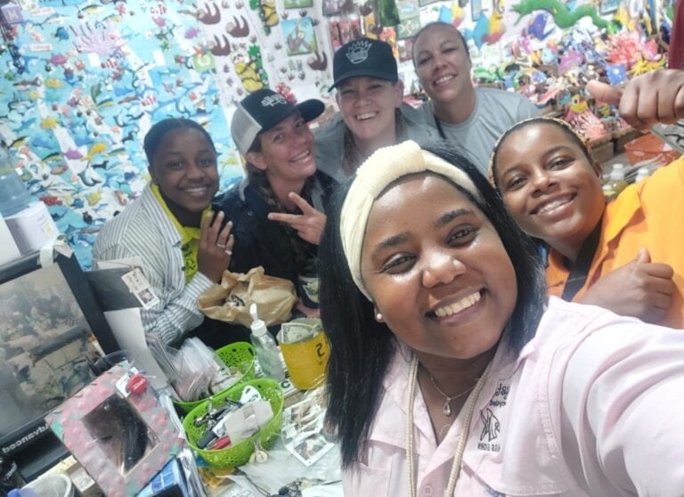 A group of smiling women posing inside a colorful, ocean-themed decor store or shop in Roatan, Honduras, with various toys, souvenirs, and supplies visible on the table and shelves behind them.
