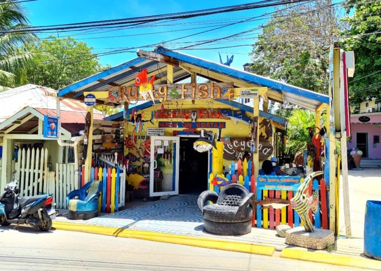 A colorful fishing shop with a bright yellow facade and a blue roof is located along a street in Roatan, Honduras, featuring fishing-themed decorations, a painted sign reading "Ruggy Fish" and "Made on Roatan," and a small seating area with a tire-shaped chair.