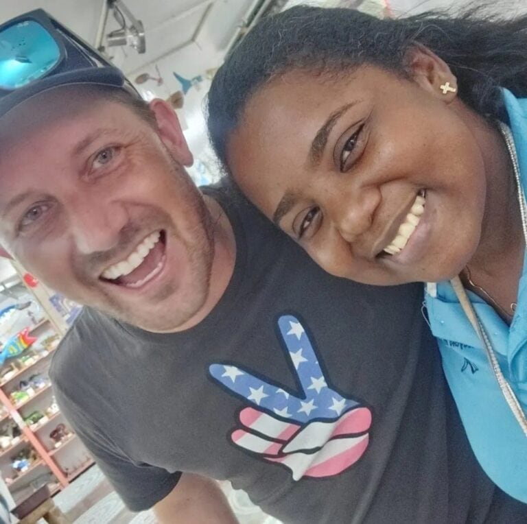 Two smiling individuals, a man wearing a black T-shirt with a patriotic peace sign and sunglasses on his head, and a woman in a blue jacket with cross earrings, pose closely together inside a shop on Roatan, Bay Islands, Honduras.