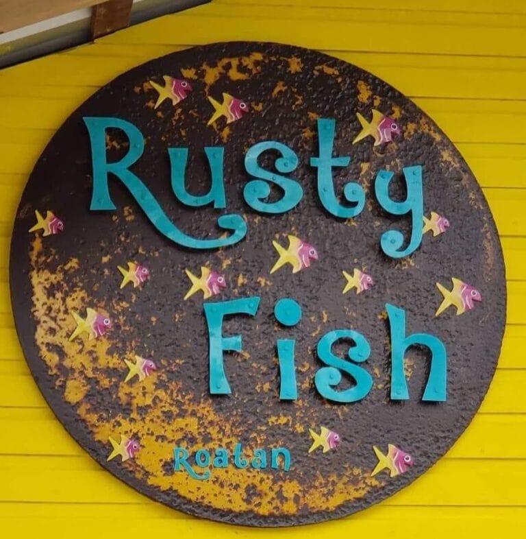 A round, rusted metal sign with the words "Rusty Fish" and "Roatan" in blue letters, decorated with small colorful fish illustrations, is mounted on a bright yellow wall in Roatan, Honduras.
