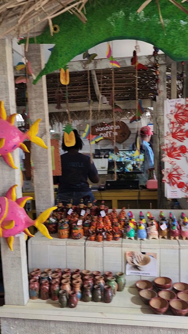 A colorful market stall in Roatan, Honduras, displays handcrafted pottery, decorative figurines, and vibrant ornaments with tropical-themed decorations hanging from the ceiling.