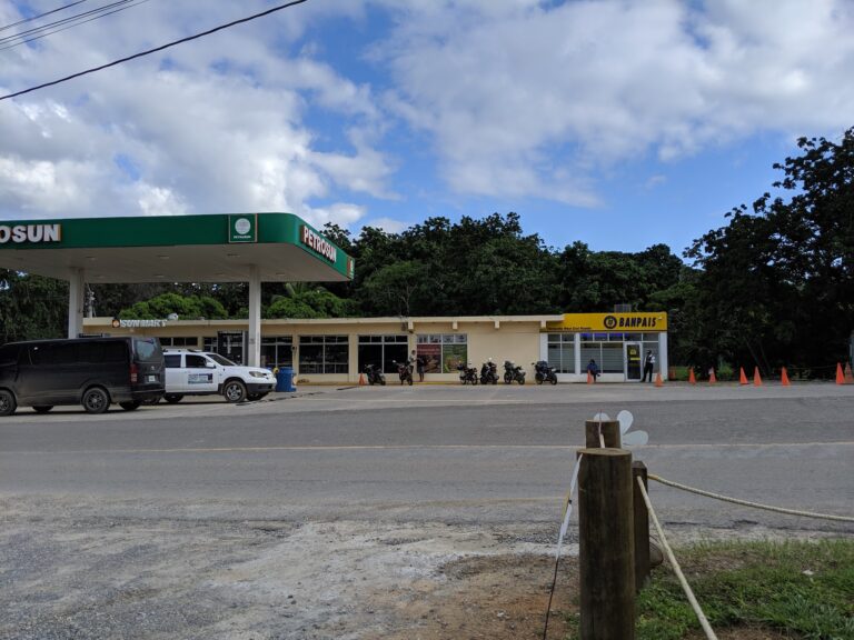 A Petrosun gas station and Sun Mart convenience store are situated along a street in Roatan, Honduras, with a background of lush green trees and a partly cloudy blue sky.