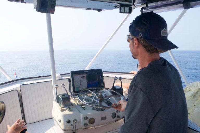 A man in a baseball cap and sunglasses operates a boat's steering and navigation system on the open ocean near Roatan, Honduras.